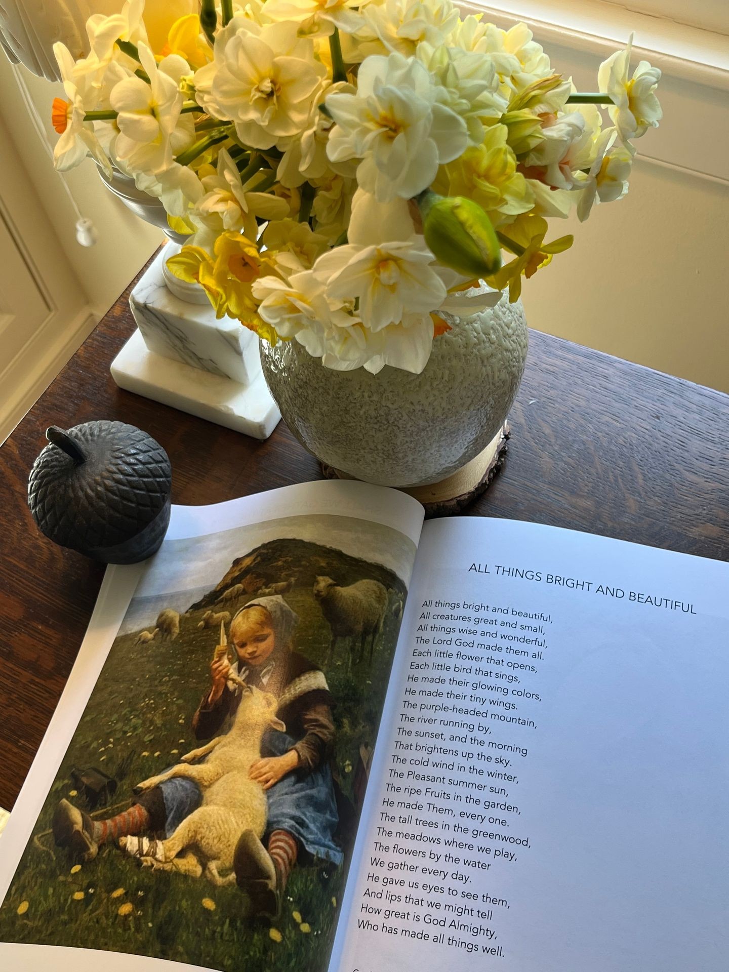 A book of Hymns for people living with Alzheimer’s dementia, featuring a little girl feeding a lamb. There is an alabaster lamp and a vase full of daffodils and jonquils. Memory care books for people living with dementia.