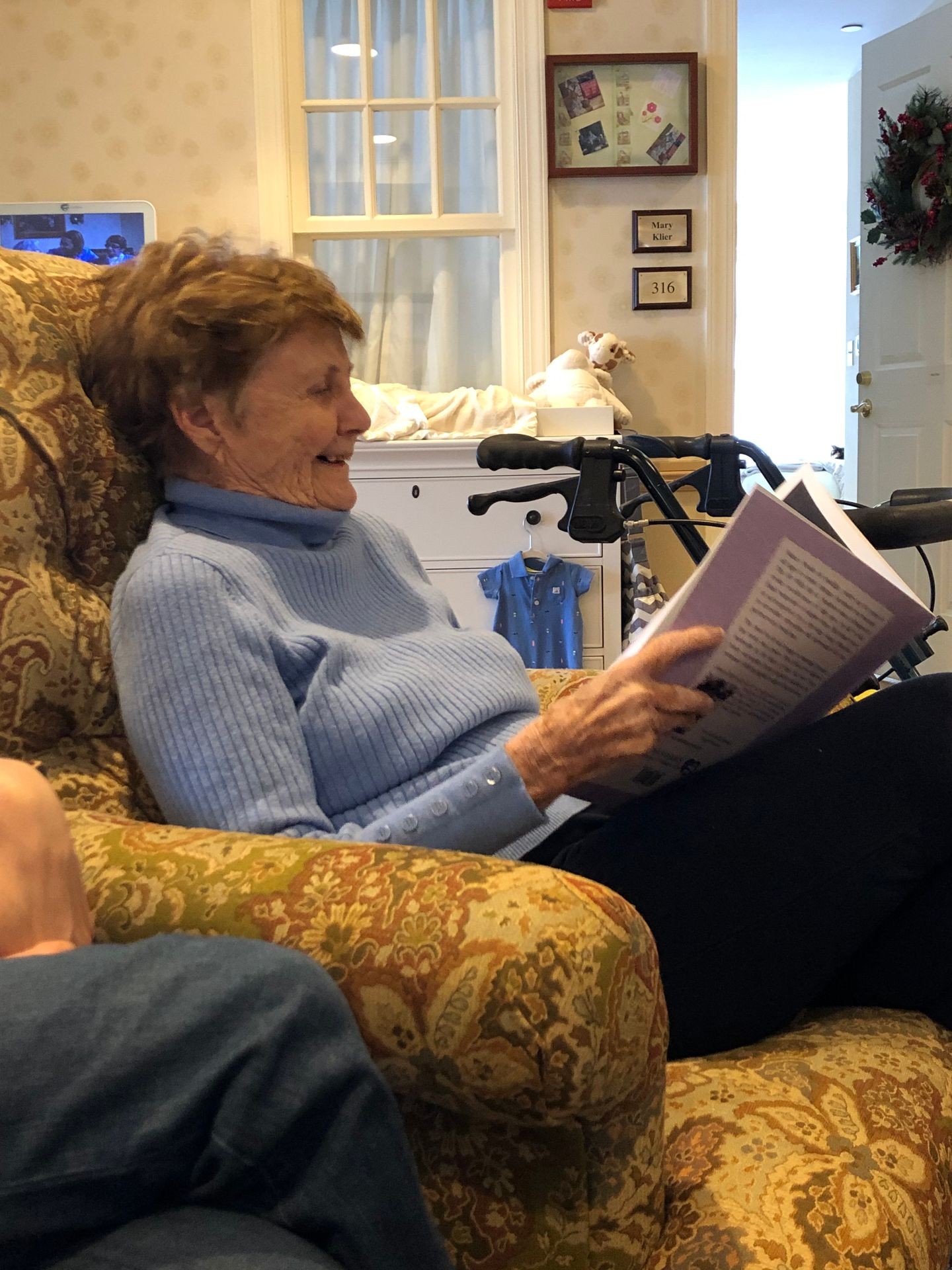 An elder woman sits and smiles as she reads a nostalgic poetry book, formatted just for her. Her walker is to one side and she is wearing a powder blue sweater with buttons down the sleeves.  