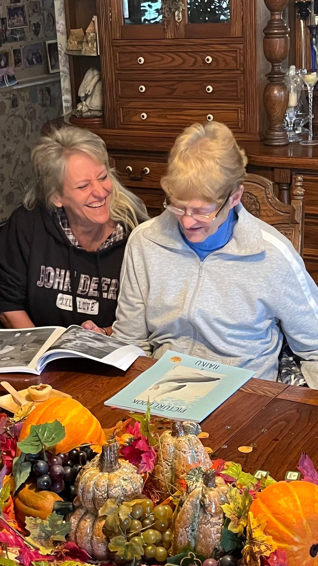 A smiling woman with a sweatsuit and glasses reads a book with her niece who is laughing. The are reading a book of Haiku. NANA’S BOOKS are for people living with dementia.