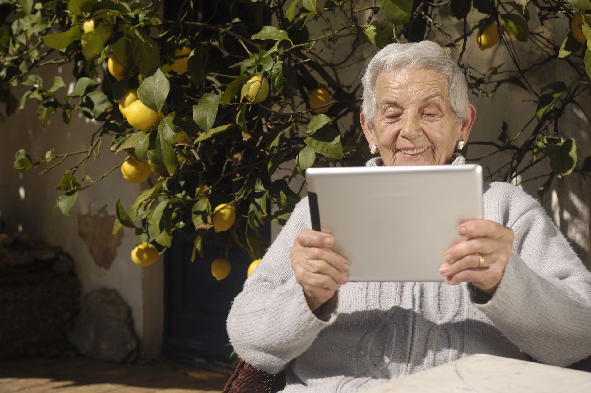  Smiling older woman sitting next to a lemon tree and reading on a tablet. She has on a tan sweater. NANA’S BOOKS help her to read independently, as they are considerately-formatted, large print books which support reminiscence. 